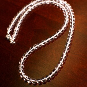May include: A clear crystal beaded necklace with a silver clasp. The necklace is laid out on a dark brown wooden surface.