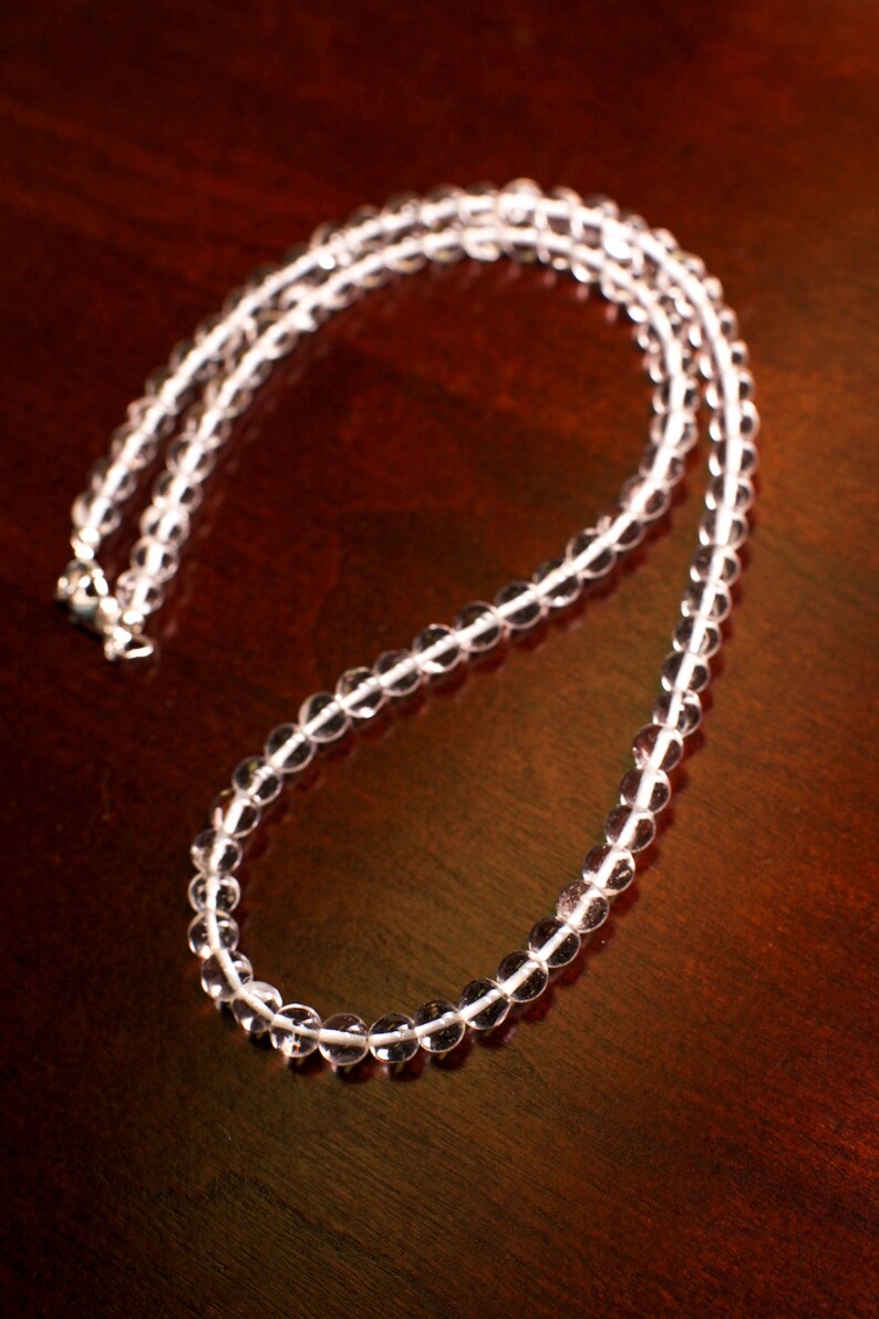 May include: A clear crystal beaded necklace with a silver clasp. The necklace is laid out on a dark brown wooden surface.