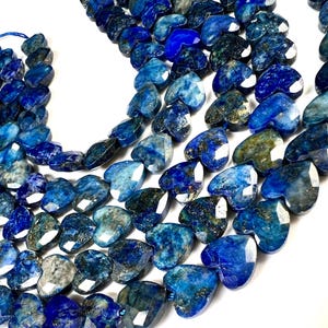 May include: Strands of heart-shaped lapis lazuli beads in varying shades of blue. The beads are faceted, catching the light and revealing a mix of dark and light blue hues. The beads are strung together with blue string.
