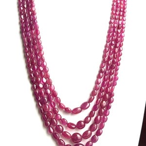 May include: A multi-strand necklace featuring oval-shaped, deep pink gemstones. The necklace is displayed on a white surface, showcasing the vibrant color and polished texture of the stones. The necklace has a gold clasp.