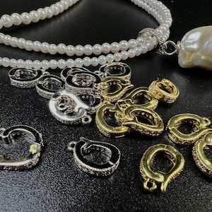 Könnte beinhalten: Eine Sammlung von Schmuckteilen, darunter gold- und silberfarbene Verschlüsse mit klaren Kristallakzenten. Eine weiße Perlenkette mit einem großen Barockperlenanhänger ist ebenfalls vorhanden. Die Artikel sind auf einer schwarzen Oberfläche angeordnet.