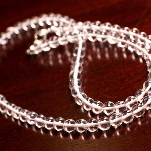 May include: A clear quartz crystal beaded necklace with a silver clasp. The necklace is laid out on a dark brown surface.