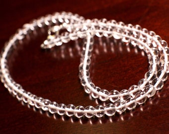 May include: A clear quartz crystal beaded necklace with a silver clasp. The necklace is laid out on a dark brown surface.