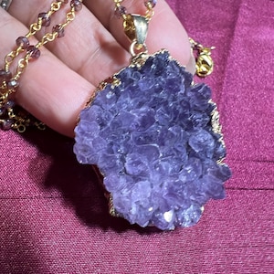 May include: A purple amethyst pendant necklace with a gold-colored chain and accents. The amethyst is a rough, irregular shape with a gold-colored trim. The necklace is held against a dark pink background.