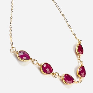 May include: A gold chain necklace featuring four teardrop-shaped, faceted deep pink ruby gemstones. Each gemstone is individually bezel-set in gold and connected by thin gold wire links, creating a delicate and symmetrical arrangement.