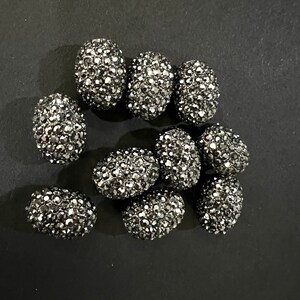 May include: A group of ten oval-shaped black beads with a sparkly, faceted finish. The beads are scattered on a black surface.