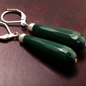 May include: A pair of elegant dangle earrings. Each earring features a faceted, dark green, teardrop-shaped bead, accented with a small white pearl and silver-colored leverback closures. The earrings are displayed on a dark brown surface.
