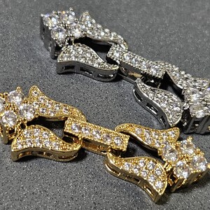 May include: Two iced-out chain links, one gold and one silver, are displayed on a gray surface. Both links are encrusted with numerous small, clear gemstones, creating a sparkling effect. The gold link is on the bottom.