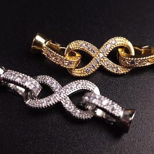 May include: Two infinity-shaped clasps for jewelry, one gold and one silver, both with clear cubic zirconia stones.