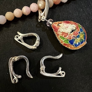 Könnte beinhalten: Vier silberfarbene Ohrringverschlüsse aus Metall, zwei sind offen und zwei sind geschlossen. Eine rosa-weiße Perlenkette mit einem silberfarbenen Metallverschluss und einem bunten Emaille-Anhänger hängt über den Verschlüssen.