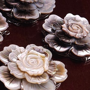 May include: Three iridescent mother-of-pearl flower charms with a rose design. The flowers are layered and have a delicate, intricate design. The charms are attached to silver rings.