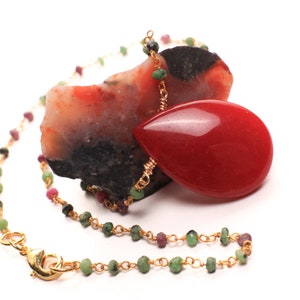 Puede incluir: Un collar de cadena dorada con cuentas verdes y rosas y un colgante rojo en forma de lágrima. El colgante descansa sobre una piedra rugosa roja y negra.