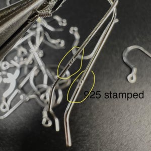 May include: Close-up of silver-colored earring hooks with a 925 stamp, indicating sterling silver. The hooks are in various stages of being formed, with a pair of pliers visible in the background. The text "925 stamped" is also visible.