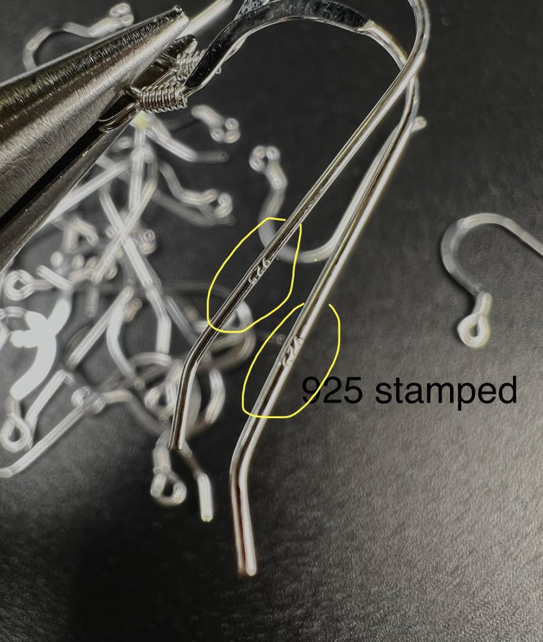 May include: Close-up of silver-colored earring hooks with a 925 stamp, indicating sterling silver. The hooks are in various stages of being formed, with a pair of pliers visible in the background. The text "925 stamped" is also visible.