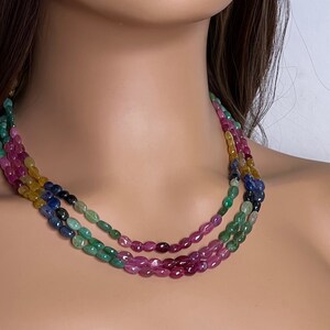 May include: A multi-strand necklace made of oval-shaped gemstones in shades of pink, green, blue, and yellow. The necklace is layered with the pink gemstones on the bottom, followed by green, blue, and yellow.
