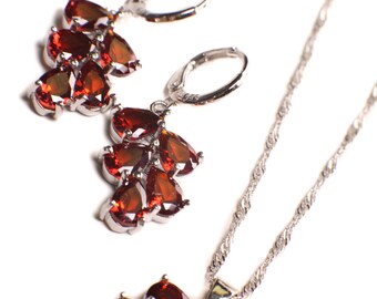 May include: A silver necklace and earrings set with a cluster of red gemstone pendants. The necklace has a delicate chain and the earrings have small hoop closures.