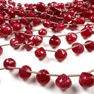 May include: Strands of faceted, deep red gemstone beads. The beads are strung on thin, gold-colored wire, creating a rich, textured appearance. The gemstones have a polished, reflective surface, and are arranged in a close-up shot.