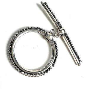 May include: Silver toggle clasp with a rope-like design. The clasp is made of two pieces, a round ring and a straight bar that slides through the ring.