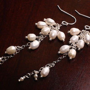 Peut inclure: Une paire de boucles d'oreilles pendantes en argent avec des perles blanches. Les boucles d'oreilles ont plusieurs brins de perles qui cascadent du haut.