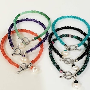 May include: Five layered beaded bracelets with toggle clasps. The bracelets are made of green, purple, black, turquoise, and pink beads. Each bracelet has a white pearl charm.