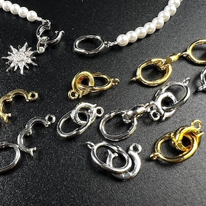 Könnte beinhalten: Eine Auswahl an Schmuckverschlüssen und -verbindern in Gold- und Silbertönen. Die Verschlüsse variieren in Form und Design, einschließlich ovaler, runder und dekorativer Stile. Eine Perlenkette ist ebenfalls sichtbar, zusammen mit einem sonnenförmigen Anhänger.