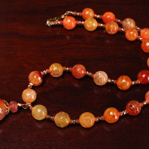 May include: A necklace made of orange and yellow agate beads with a large orange agate pendant. The necklace is on a brown wooden surface.