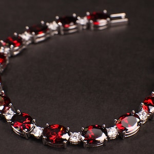 May include: A silver bracelet with a row of oval-shaped red gemstones and smaller clear gemstones. The bracelet is on a black background.