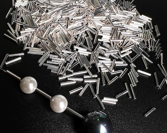 Può includere: Primo piano di un mucchio di tubi metallici argentati, sparsi su una superficie nera. Una collana con tre perle, due bianche e una nera, è parzialmente visibile in primo piano.