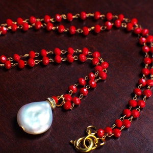 May include: A red beaded necklace with a large white pearl pendant. The necklace is made of small, faceted red beads and a gold chain.