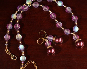 May include: A set of iridescent purple and gold necklace and earrings. The necklace is made of round beads with a pearlescent finish. The earrings are made of a single round bead with a pearlescent finish and a smaller round bead with a dark purple finish.