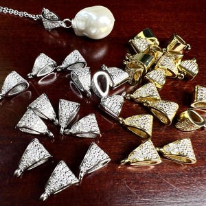 Pode incluir: Uma coleção de achados de joalheria, incluindo pingentes prateados e dourados, e um colar com pingente de pérola. Os pingentes são adornados com pequenas pedras preciosas transparentes. O pingente de pérola é grande e esbranquiçado.