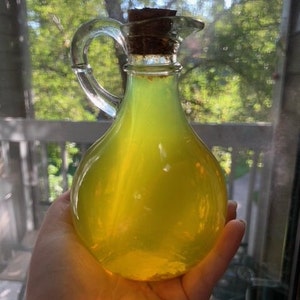 May include: A clear glass decanter with a handle and spout, filled with a bright yellow liquid. The decanter is sealed with a cork stopper. The background shows a blurred view of a window and trees.