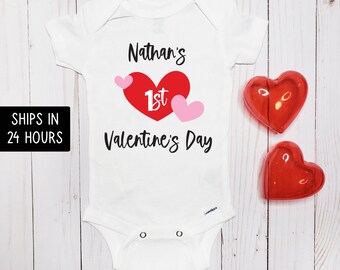 May include: White baby onesie with the text "Nathan's 1st Valentine's Day" in black script. A large red heart with "1st" in white and two pink hearts are printed on the front. Two red heart-shaped decorations are next to the onesie.
