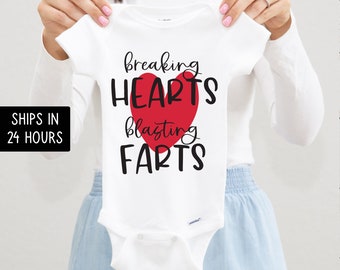 May include: White baby onesie with the text "breaking HEARTS blasting FARTS" in black font, with a red heart graphic. The onesie is held up by a person.