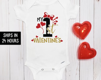 May include: White baby onesie with the text "MY 1st VALENTINE'S" in black, gold, and red. The design includes a red bow, hearts, and the number "1". Two red heart-shaped decorations are on the right.