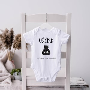 Könnte beinhalten: Weißer Baby-Body mit schwarzem Text und einer Grafik eines Geldsacks. Der Text auf dem Body lautet "UŚCISK" über der Geldsack-Grafik mit "20$" darin. Darunter steht der Text "*STUDIA SA DROGIE". Der Body hängt an einem weißen Kleiderbügel.