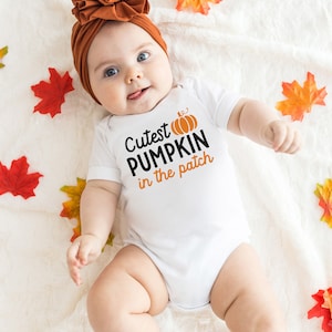 May include: A baby wearing a white onesie with the text "Cutest PUMPKIN in the patch" and an orange pumpkin graphic. The baby is also wearing a rust-colored headband. Fall leaves are scattered around the baby.