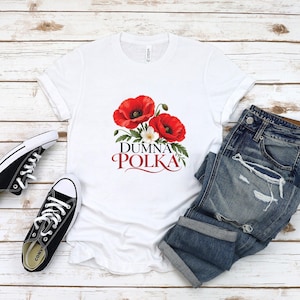 May include: A white short-sleeved t-shirt featuring a graphic of two large red poppy flowers with green leaves and a small white flower. Below the floral design, the text reads DUMNA POLKA in black and red lettering.