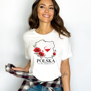 May include: A white short-sleeved t-shirt featuring a black outline map of Poland, red poppy flowers, and a heart with the Polish flag colors. Below the graphic, the text reads POLSKA W SERCU in black capital letters.