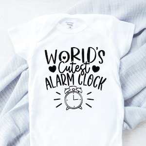 May include: White baby onesie with the text "WORLD'S Cutest ALARM CLOCK" in black. The design includes two heart shapes and a graphic of an alarm clock. The onesie is made of soft fabric and is designed for infants.