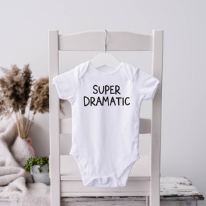 May include: A white baby onesie with the words "SUPER DRAMATIC" printed in black. The onesie is hanging on a white hanger, which is suspended from a white chair. The background includes a vase with dried plants and a blanket.