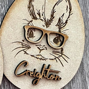 May include: Wooden Easter egg ornament with a laser-cut design of a bunny wearing glasses. The bunny's name, "Creighton", is written below the image. The ornament is a natural wood color.