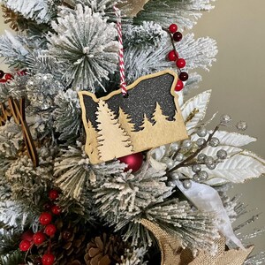 May include: A wooden Christmas ornament in the shape of the state of Oregon, featuring a silhouette of trees against a glittery black background. The ornament hangs from a red and white striped string, displayed on a snow-covered Christmas tree.