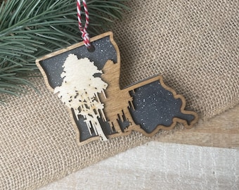 May include: A wooden Christmas ornament shaped like the state of Louisiana with a silhouette of a tree inside. The ornament is painted black with white glitter and has a red and white twine hanger.