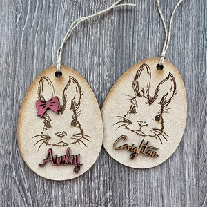 May include: Two wooden Easter egg ornaments with bunny faces, one with a pink bow and the name "Ainsley" and the other with the name "Creighton".