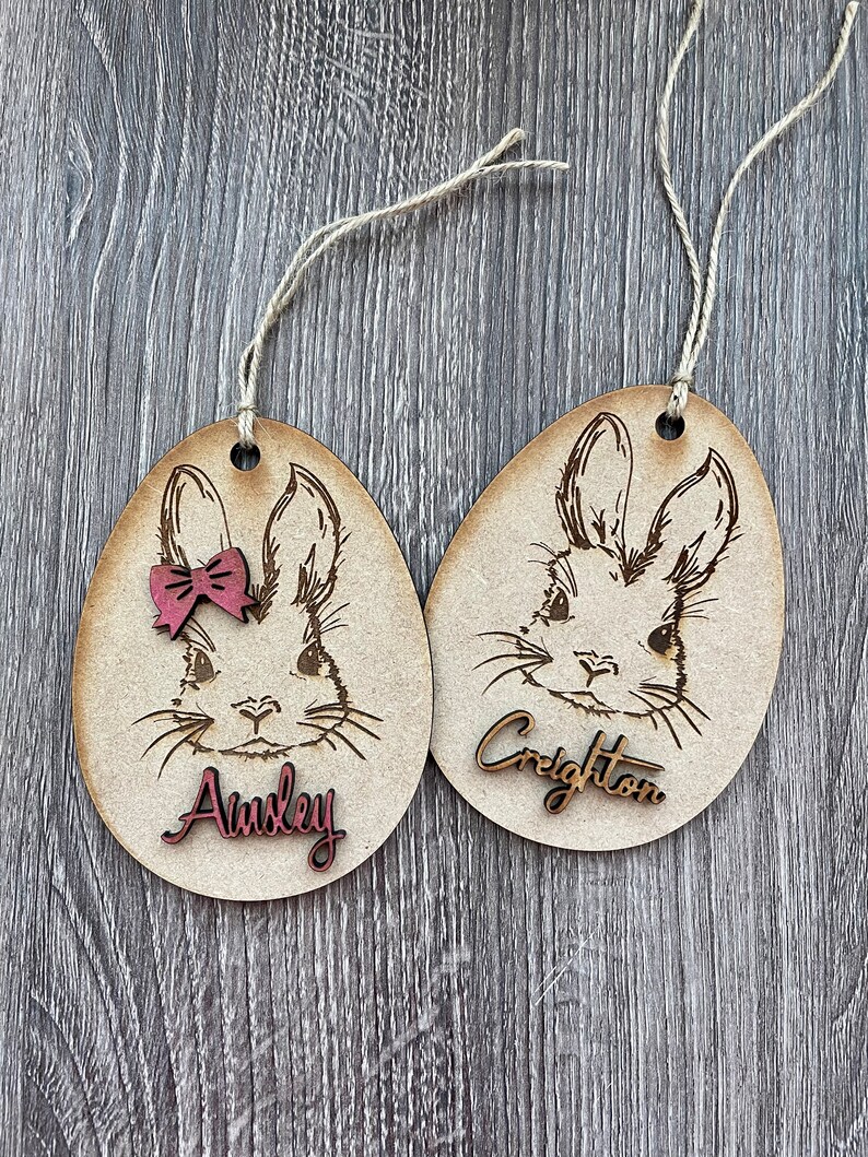 May include: Two wooden Easter egg ornaments with bunny faces, one with a pink bow and the name "Ainsley" and the other with the name "Creighton".