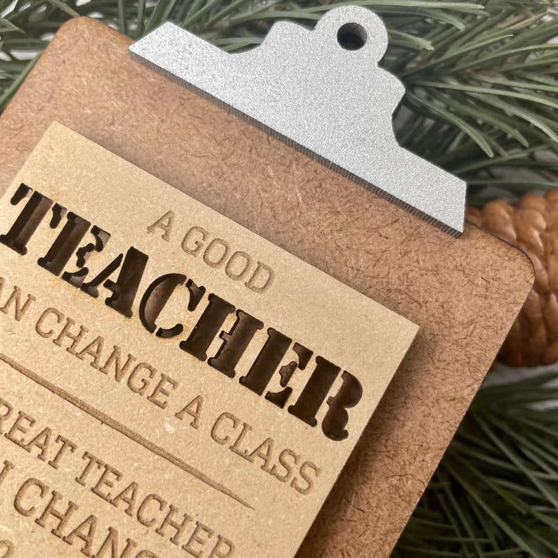 Pode incluir: Um ornamento de madeira em forma de prancheta com uma nota de papel castanho que diz "Um bom professor pode mudar uma turma". A nota est&aacute; presa &agrave; prancheta com um clipe de metal prateado.