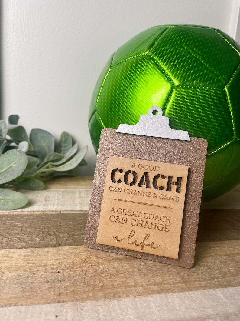 May include: A wooden sign with a clipboard design and the text "A good coach can change a game. A great coach can change a life."