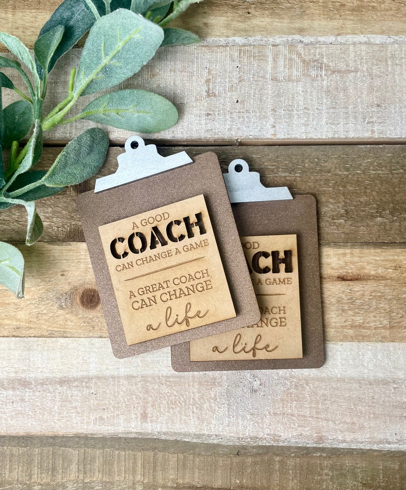 May include: Two wooden clipboard-shaped signs with the text "A good coach can change a game. A great coach can change a life." The signs are brown with white lettering and are sitting on a wooden surface.