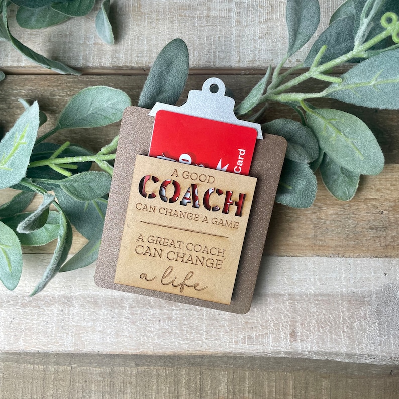 May include: A wooden gift card holder with a cutout design that reads "A good coach can change a game. A great coach can change a life." The holder has a slot for a gift card and a silver clip.